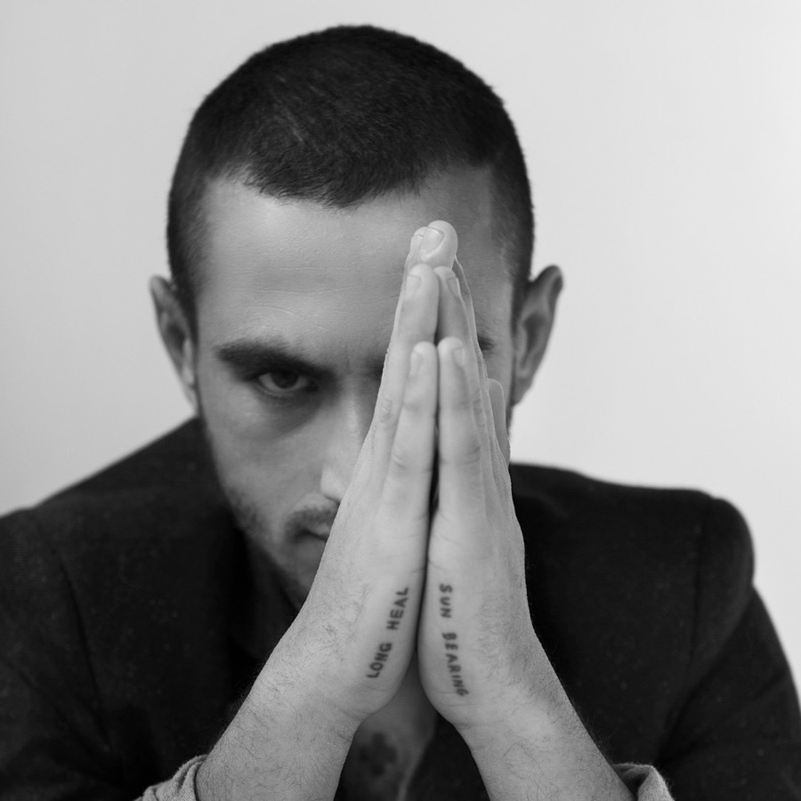 Nabil Sabio Azadi in black and white, seated at a table in a dark jacket, hands pressed together upright in front of the face so that one eye looks out past them. Along the edges of the hands are tattooed the words Long heal and Sun bearing.