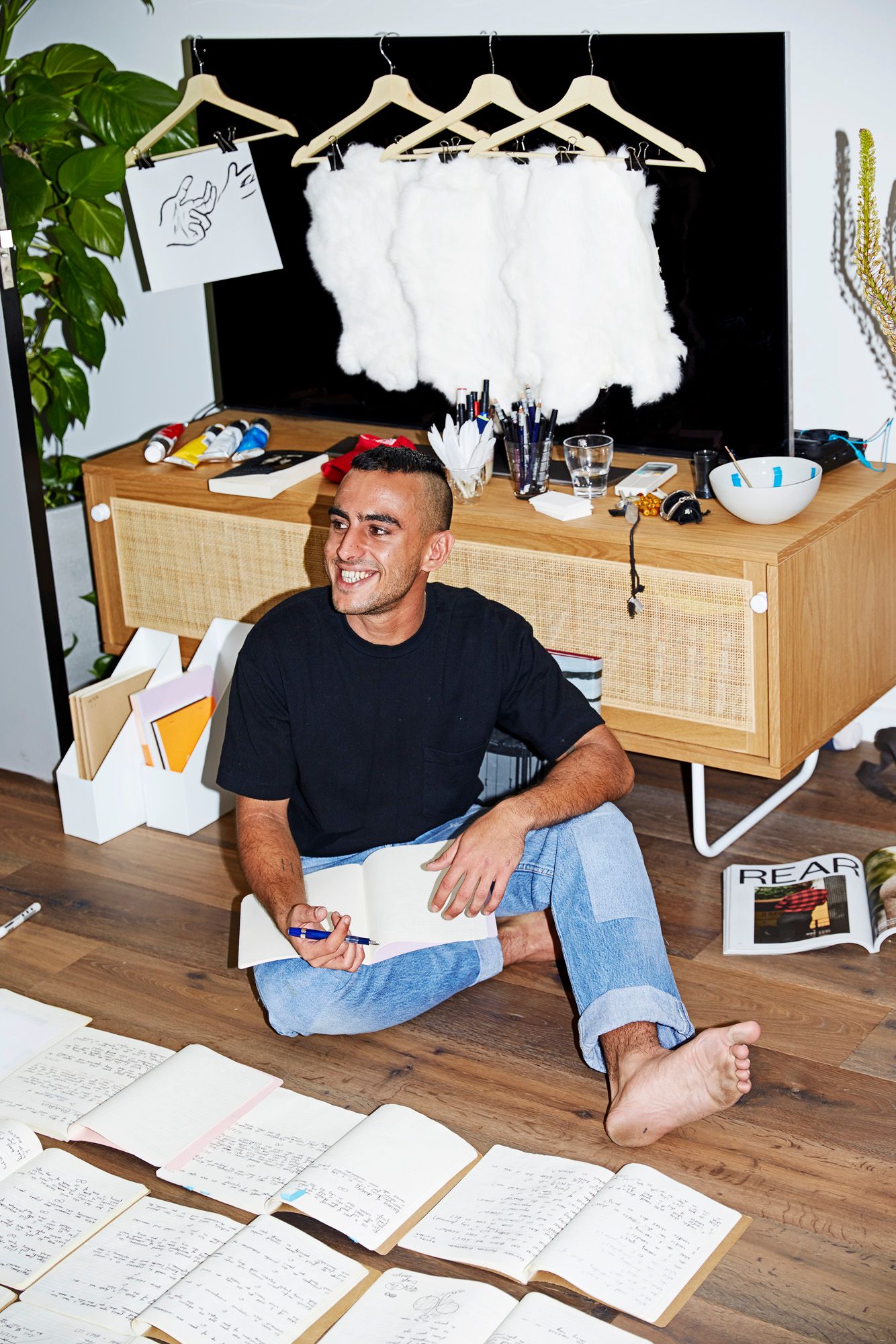 Nabil Sabio Azadi sitting on the floor of a temporary studio in a black T-shirt and jeans, smiling, a notebook open on one knee and open notebooks laid out on the floor before him. Above, four white pelts and a drawing of a hand hang from coat hangers.